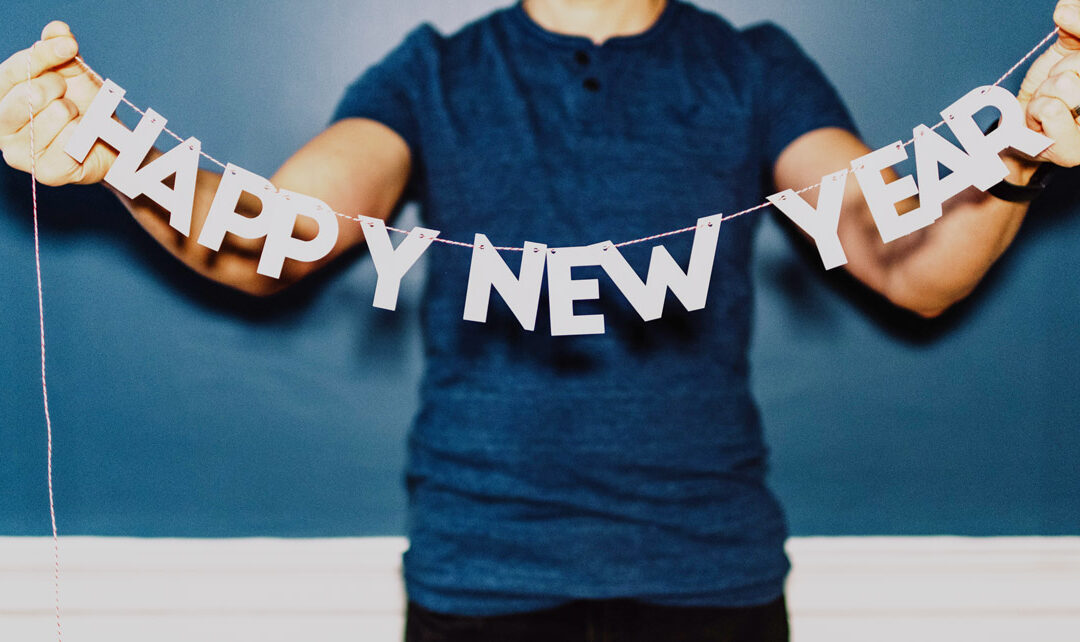 A person in a blue shirt standing against a blue wall holds up a string of paper letters that spell out Happy New Year